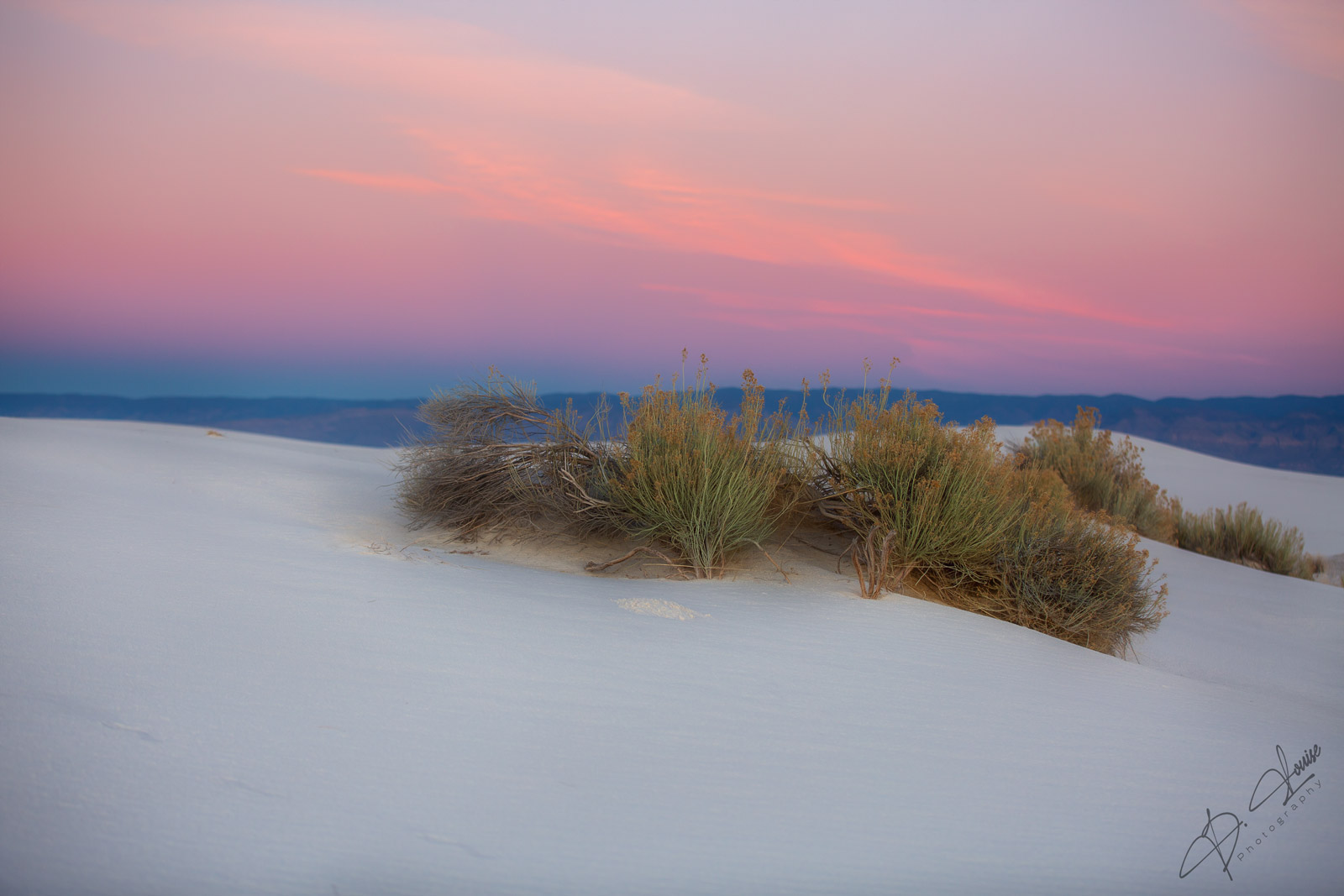 The pure white gypsum’s reflective glow of White Sands National Park glows as the last bit of sun paints the sky in a pink...